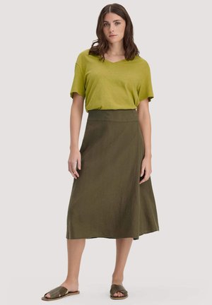 Vrouw met schouderlang bruin haar, draagt een losse groene top met korte mouwen, een olijfgroene rok met hoge taille en groene slippers, staand in een neutrale houding.