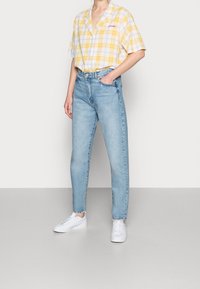 Kortärmad gul och vit rutig skjorta i kombination med ljusblå högmidjade jeans. Vita sneakers. Enfärgad bakgrund.