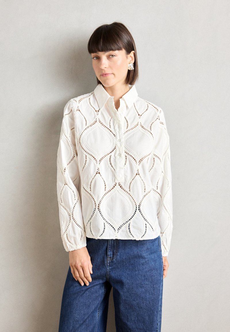 Blouse blanche avec des motifs découpés élaborés, un col, des manches longues et une coupe décontractée. Associée à un pantalon en denim bleu.