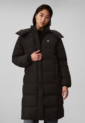 Femme portant un long manteau noir matelassé avec un col montant et le logo Calvin Klein, debout avec une main près de la fermeture éclair.