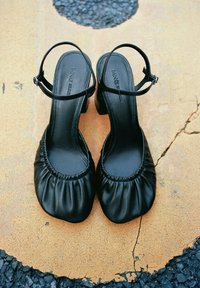 Černé kožené lodičky na podpatku se zavazováním na patě a stahovaným vrškem položené na popraskané béžovo-černé vzorované zemi.