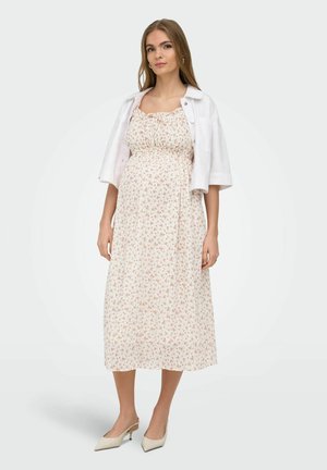 Mladá žena v bielych kvetovaných midi šatách s bielou krátkou bundou a béžovými lodičkami s nízkym podpätkom stojí pred jednofarebným pozadím.