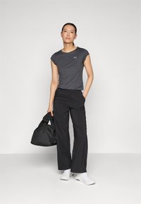 Grå t-skjorte med kort erme og logo, sammen med svarte vide bukser og hvite joggesko. Personen holder en svart treningsbag.