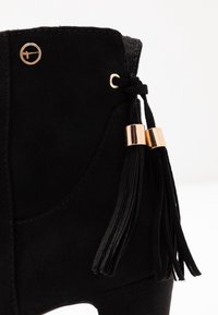 Zwarte suède enkellaars met gouden hardware, zijfranjes en een gestructureerde bovenkant. Bevat een ronde metalen accenten nabij de vetergaatjes.