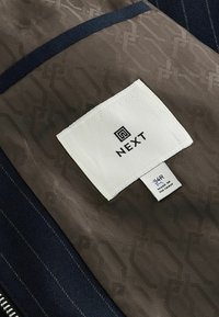 Brun stofsektion med tekstureret mønster, marineblåt stof og et hvidt mærke med "NEXT." Mærket inkluderer størrelse og "MADE IN VIETNAM."