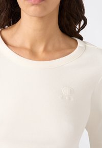 Frau trägt ein weißes, geripptes Rundhalsshirt mit einem dezenten Champion-Logo, das auf der linken Brust aufgestickt ist.
