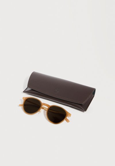 Στρογγυλά γυαλιά ηλίου σε γκριζοκάστανο χρώμα με σκουρόχρωμους φακούς, τοποθετημένα δίπλα σε μια κλειστή θήκη γυαλιών από σκουρό καφέ δέρμα, σε ανοιχτόφωρο φόντο.