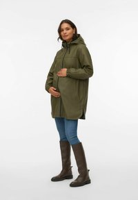 Wodoodporny płaszcz przeciwdeszczowy w oliwkowej zieleni z kapturem, zamkiem na suwak i elastycznymi mankietami. Połączony z niebieskimi dżinsami i brązowymi kozakami do kolan.