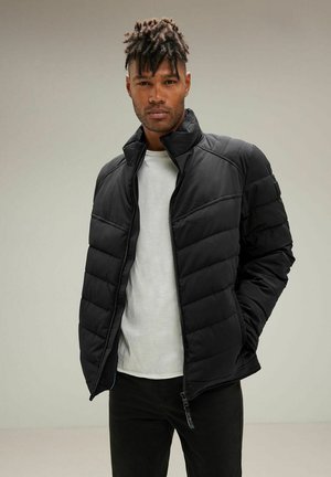 Chaqueta acolchada negra con cuello alto y cremallera. Presenta un acolchado horizontal, un corte ajustado y está fabricada en un material suave y ligero.