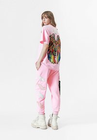 Camiseta rosa de gran tamaño con bordado colorido de alas en la parte posterior, combinada con pantalones a juego que presentan diseños de alas blancas. Botas blancas.