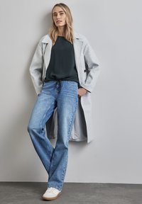 Grauer Wollmantel, dunkelgrünes Oberteil, hellblaue Jeans und weiße Sneakers. Das Outfit hat einen entspannten Schnitt und einen Gummizug in der Taille.