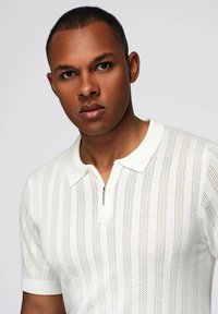 Camisa polo blanca de manga corta con patrones de punto vertical, cuello con cremallera y acabado texturizado. Diseño ligero y ajustado.