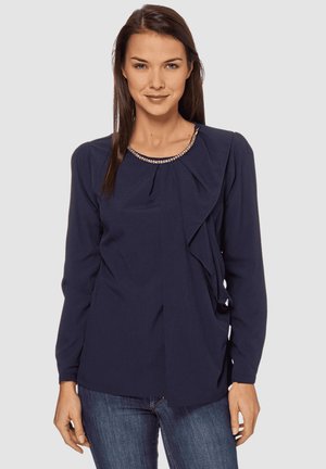 Mujer con una blusa de manga larga azul marino con cuello adornado con cuentas y detalle de volantes, combinada con jeans azules, de pie contra un fondo liso.