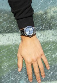 Reloj de acero inoxidable plateado en una mano, con una esfera azul, acentos plateados, display de fecha y una camisa de manga larga negra; fondo de vidrio agrietado.