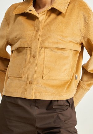 Veste en velours côtelé couleur beige avec grandes poches à rabat et fermeture par boutons, portée sur un pantalon marron foncé par une personne les mains derrière le dos.