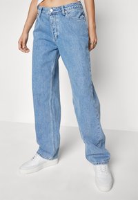 Jean large bleu clair en denim. Possède une taille haute, cinq poches et une fermeture à boutons. Porté avec des baskets blanches.