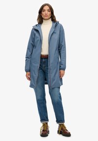 Wodoodporna niebieska kurtka z kapturem, z przodu zapinana na zamek błyskawiczny, z elastycznymi mankietami i jasnobrązowymi akcentami, w połączeniu z podwiniętymi dżinsami i brązowymi butami.