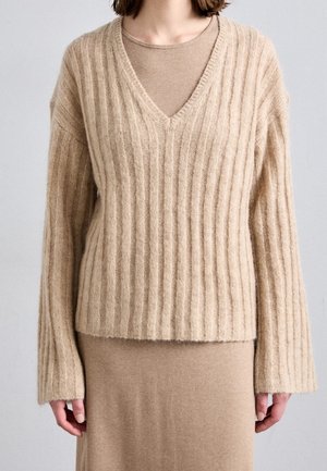 Vrouw draagt een beige geribbelde V-hals trui over een bijpassende top met lange mouwen en rok, staand voor een effen achtergrond.