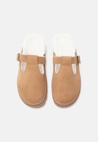 Barna velúr, slip-on papucs fehér bolyhos béléssel, széles pánttal az elülső részen és nyitott orrú kialakítással.