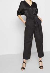 Top in satin nero con scollo a V e maniche a sbuffo, abbinato a pantaloni neri a vita alta con vita regolabile e gambe affusolate.