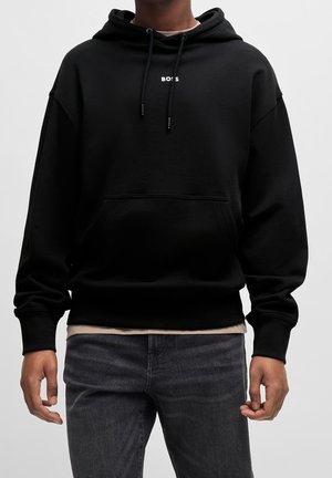 Person iført en sort hættetrøje med forlommer og "BOSS"-logo centreret på brystet, kombineret med mørke jeans og en beige undertrøje.