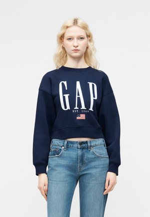 Jeune femme aux cheveux blonds portant un sweat-shirt GAP bleu marine et un jean bleu clair, debout devant un fond blanc uni.