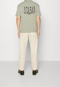 Szaro-zielona koszulka z krótkim rękawem z dużą grafiką "1981" oraz napisem "Emporio Armani Milano" na plecach, w zestawieniu z kremowymi spodniami.
