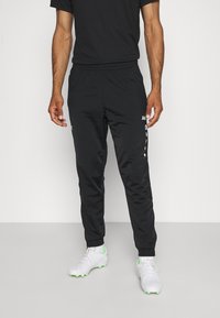 Jogging noir avec une ceinture élastique, des poches latérales et des détails en logo blanc sur la jambe gauche. Fabriqué en matériau lisse et léger.