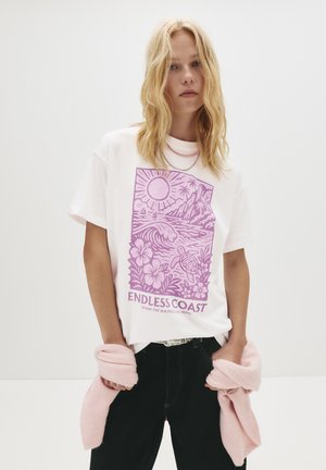 Femme blonde portant un t-shirt blanc avec un motif côtier violet et le texte "Côte sans fin", un pantalon noir et un pull rose noué autour de la taille.