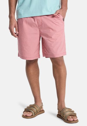 Rosa bomullsshorts med sidelommer og synlige søm-detaljer, kombinert med brune remmesandaler på en lyshudet person.