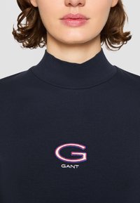 Navyblauwe coltrui van zachte stof, met een geborduurd 'G' logo in rood en wit. Strakke pasvorm met geribbelde halslijn.