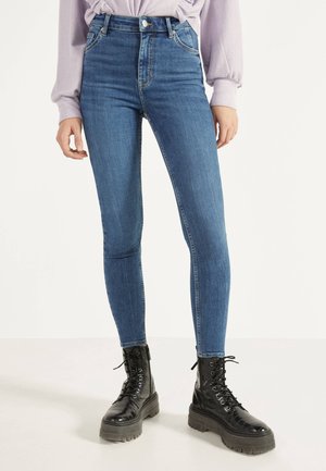 Close-up af en person iført højtaljede blå skinny jeans, en lys lilla langærmet skjorte og sorte chunky snørebåndsstøvler.