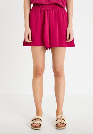 Woman wearing loose magenta shorts and beige double-strap sandals standing against a plain white background.