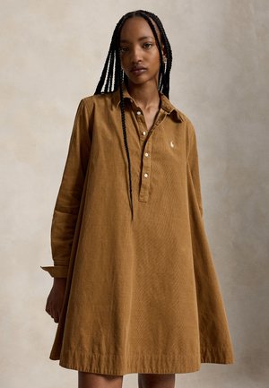 Femme aux longues tresses portant une robe marron ample à manches longues et boutonnée sur un fond neutre uni.