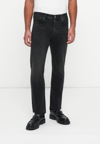 Jeans negros de pierna recta en tejido de mezclilla, con bolsillos delanteros y cierre de cremallera. Llevados con zapatos negros sin cordones, de estilo minimalista.
