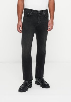 Jean noir à coupe droite en denim, avec poches avant et fermeture éclair. Porté avec des chaussures noires à enfiler, style minimaliste.