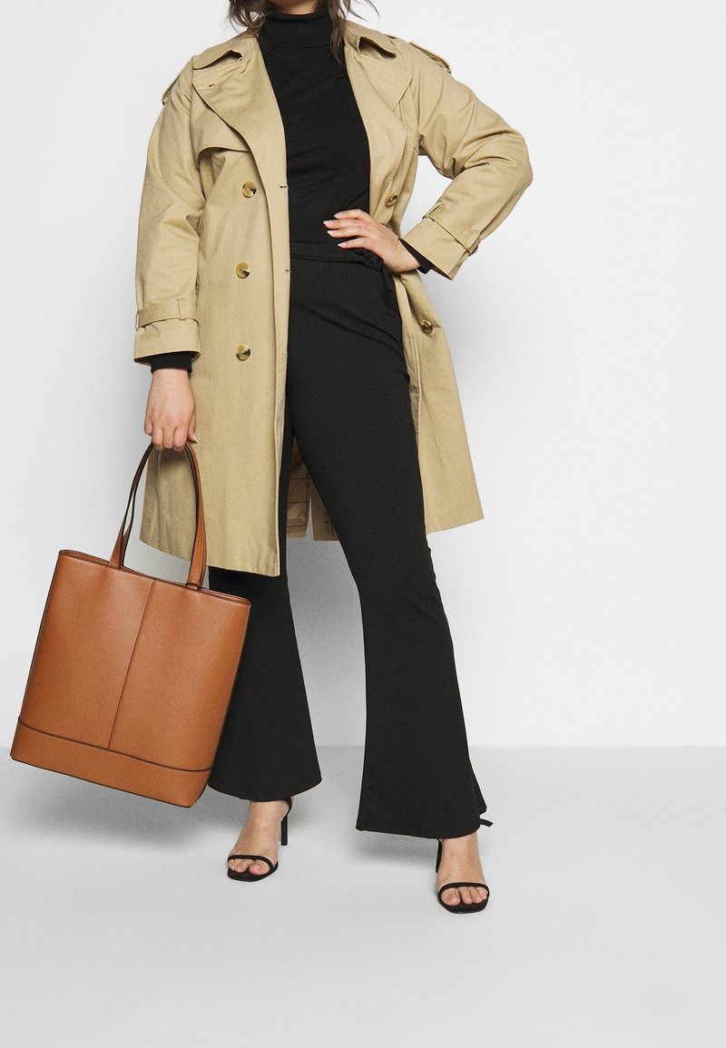 Trench beige con bottoni dorati, top nero a coste, pantaloni neri a zampa d'elefante, borsa a tracolla in pelle marrone e sandali neri con tacco.