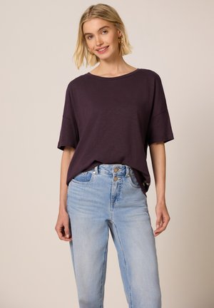 Ung kvinde iført en løs, mørkelilla T-shirt og lyseblå højttaljede jeans, smilende, stående foran en ensfarvet baggrund.