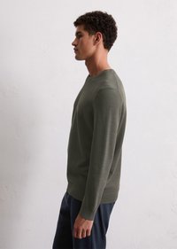 Olive green knit sweater with textured design, round neckline, and long sleeves, worn with navy pants, displayed against a light background.