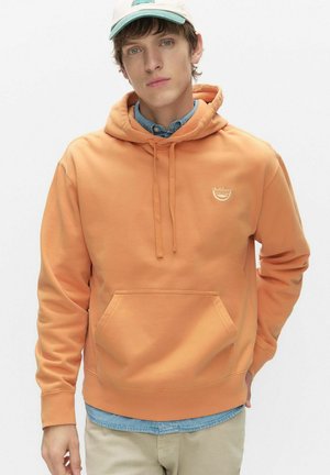 Młody mężczyzna w jasnopomarańczowej bluzie z kapturem na niebieskiej koszuli jeansowej i kremowej czapce, stojący na jednolitym tle.