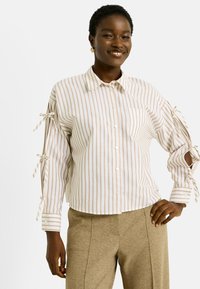 Personne souriante portant une chemise boutonnée à rayures beiges avec des détails de manches nouées et un pantalon beige, se tenant avec une main sur la hanche.