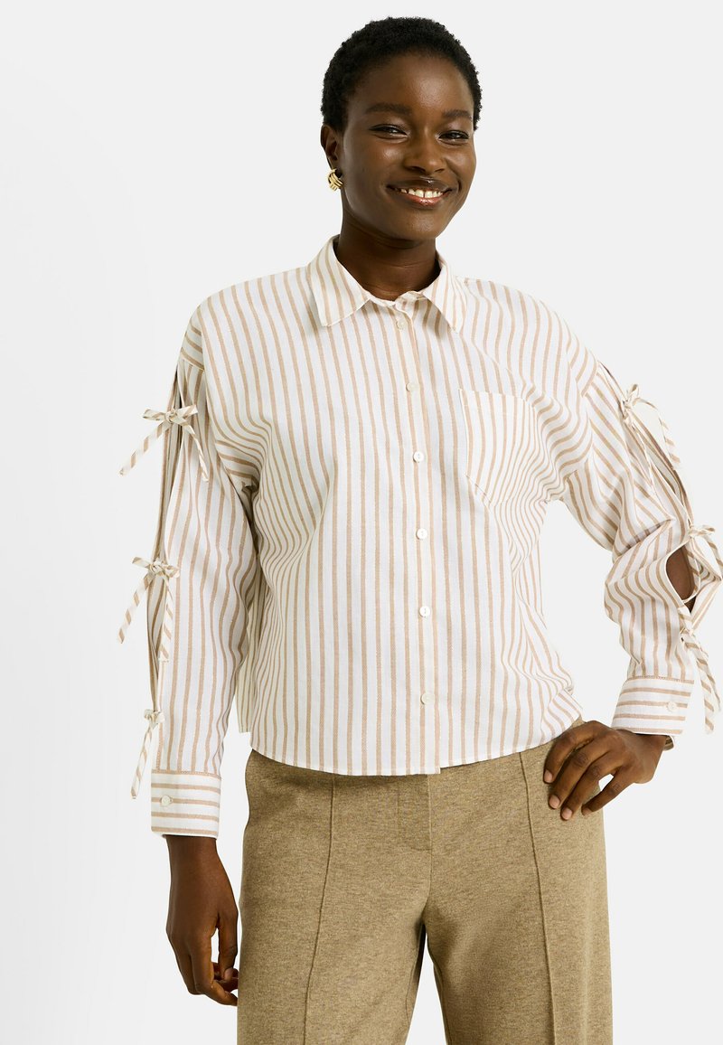 Personne souriante portant une chemise boutonnée à rayures beiges avec des détails de manches nouées et un pantalon beige, se tenant avec une main sur la hanche.