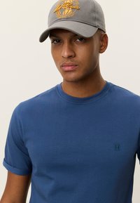 Μπλε βαμβακερό μπλουζάκι με κοντά μανίκια γυρισμένα και πράσινο κεντημένο λογότυπο "II" στο στήθος. Γκρι καπέλο με κεντημένο λογότυπο.