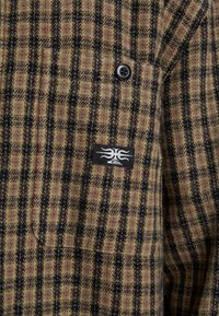 Chaqueta de lana con un patrón a cuadros en marrón y negro, con acentos rojos, un botón negro y un parche de logo en el lado izquierdo.