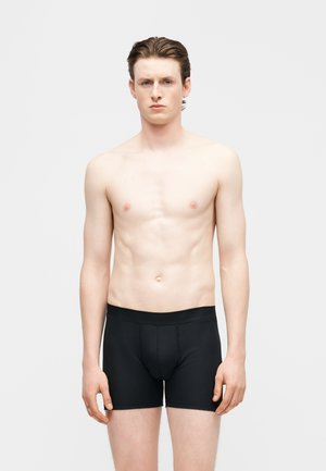 Joven de pie contra un fondo liso, vistiendo calzoncillos bóxer negros, mirando hacia adelante con expresión neutral.