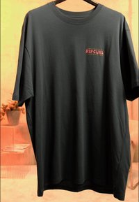 Zwart T-shirt met korte mouwen en een kleurrijk "RIP CURL" logo op de linkerborst, hangend aan een zwarte hanger in een zacht verlichte kamer.