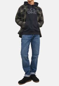 Black hoodie with gray text, plaid green and black button-up shirt, blue jeans, and black suede shoes, standing against a white background.