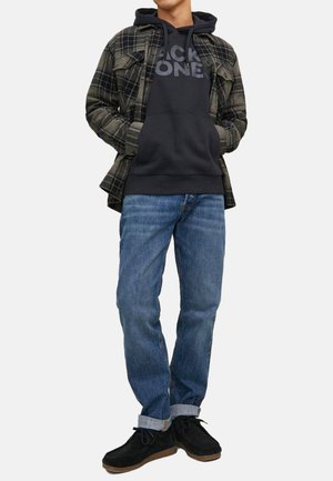 Schwarzer Hoodie mit grauem Text, karierter grün-schwarzer Hemd mit Knöpfen, blaue Jeans und schwarze Wildlederschuhe, die vor einem weißen Hintergrund stehen.
