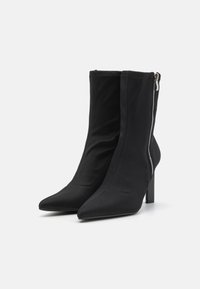 Botas de tobillo negras hechas de tela suave, con punta puntiaguda y un alto tacón cuadrado. Cierre lateral con cremallera y un acento metálico.