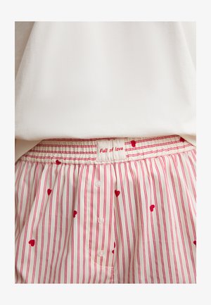 Pantalones cortos de pijama con cintura elástica, rayas verticales rojas y patrones de corazones, con una etiqueta que dice "Lleno de amor" y pequeños botones frontales.